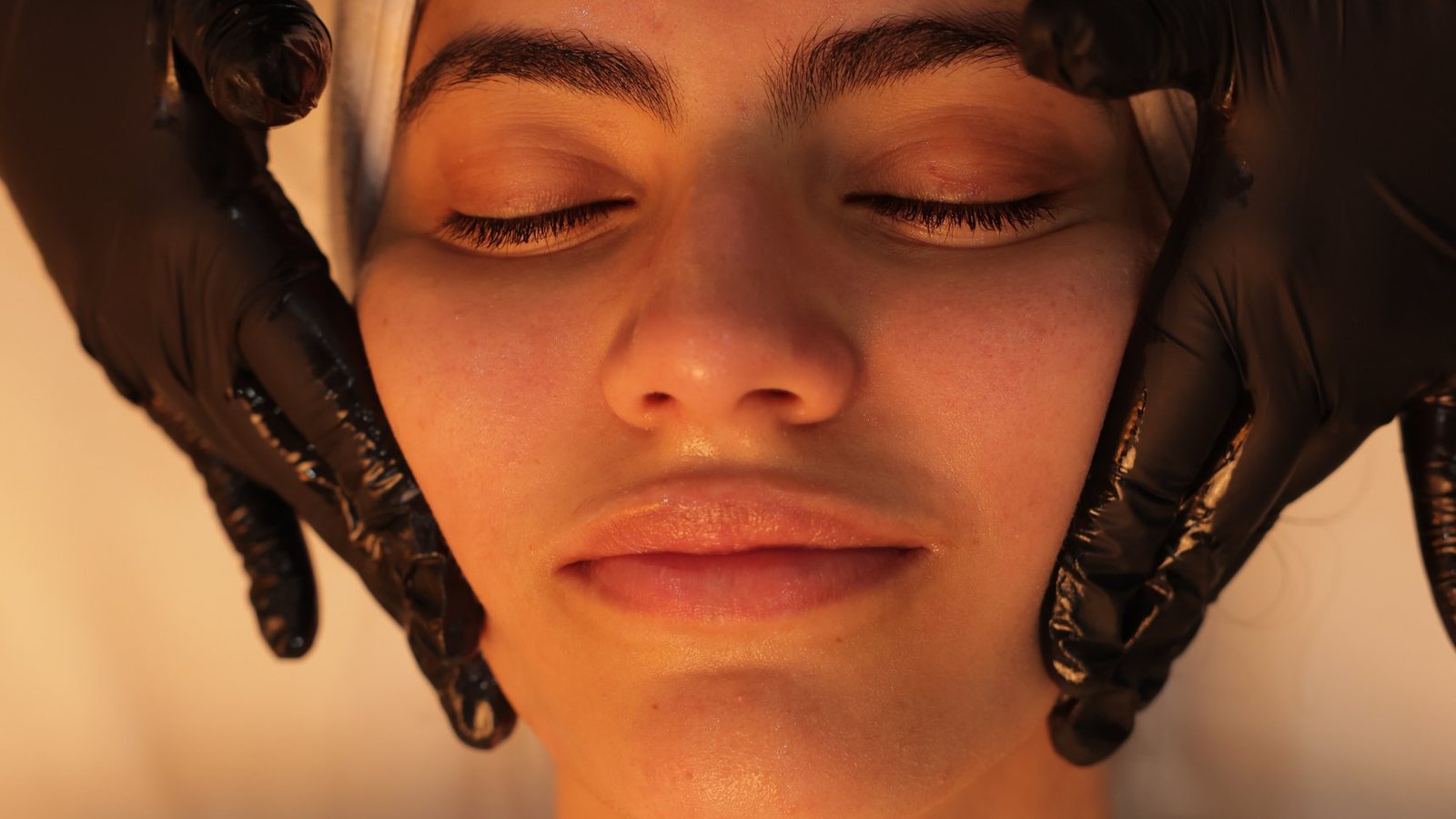 Close-up of glowing, hydrated skin during a 23MD facial, with a clinician's gloved hands framing the patient's face